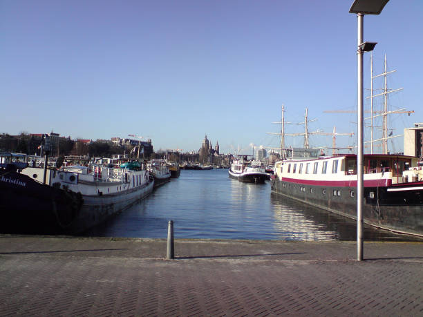This image depicts a waterfront scene, likely in a Dutch city such as Amsterdam. Several large boats are docked along both sides of a canal, with masts of ships visible in the background on the right. The clear blue sky suggests a bright day, and in the distance, a large building with towers, possibly a church or a historic structure, can be seen along the skyline. The overall atmosphere is calm, with the stillness of the water reflecting the boats and ships moored along the quay. The location has an urban harbor or marina feel, with a mix of historical and modern elements.