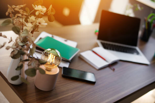 istock Warm, inviting workspace featuring a glowing light bulb, a vase, and a neatly arranged desk with a laptop, notebook, and smartphone. The soft sunlight adds to the calm and productive atmosphere. 2171020867