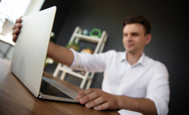 A man in a white shirt opens his laptop in a modern office setting, with a bookshelf visible in the background. The photo captures a moment of preparation and focus in a professional environment.