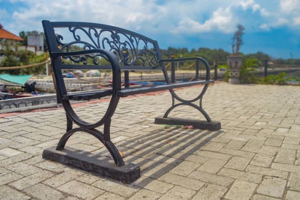 Elegant black metal bench on a paved surface with a soft focus background of blue sky, trees, and water. Perfect for themes of outdoor relaxation, park settings, and serene environments
