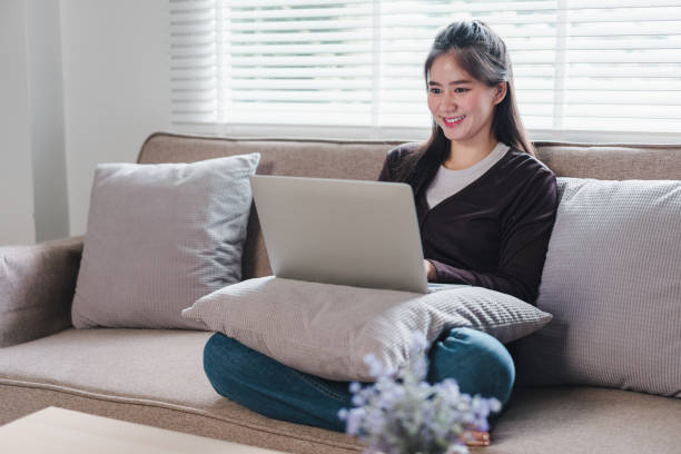 istock A young woman is sitting comfortably on sofa, using laptop while smiling. cozy atmosphere is enhanced by soft cushions and natural light streaming through window, creating warm and inviting space. 2171060011