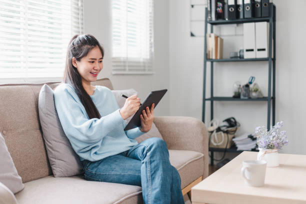 istock A young woman is sitting comfortably on sofa, smiling while using tablet. cozy living room features natural light and stylish decor, creating warm atmosphere. 2171060103