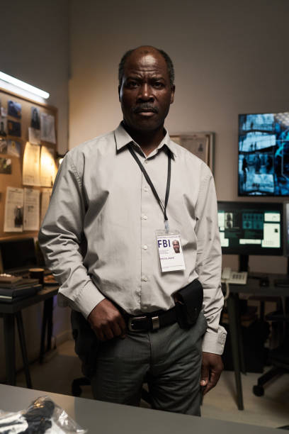 Investigating detective in well-equipped surveillance office with multiple monitors, working on evidence and files from different cases. Wearing official identification badge and gun holster