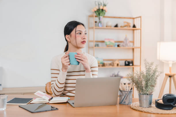 istock A young woman sits at desk, holding blue mug while looking thoughtfully away from her laptop. workspace is bright and organized, featuring plants and decorative items, creating calm atmosphere. 2171060251