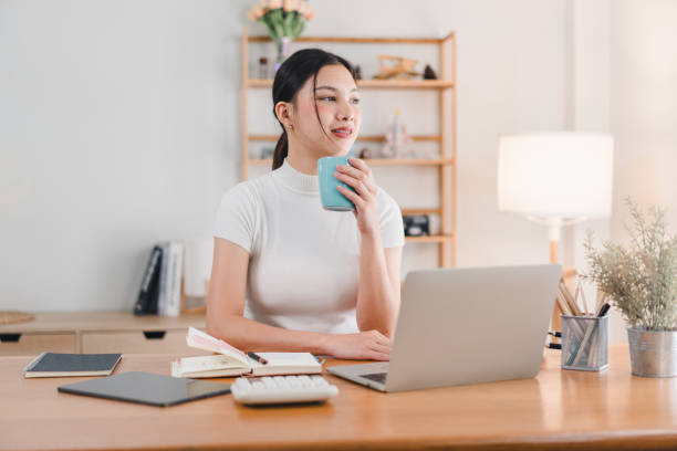 istock A young woman sits at modern desk, holding blue mug while looking thoughtfully at her laptop. workspace is bright and organized, creating calm atmosphere for productivity. 2171060287