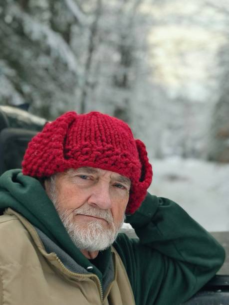 Bearded senior man, resting on his green truck(partial view of truck only),and looking with a serious expression at the camera. Heavily snowed on rural road dirt driveway and snowy woods in the background. The man is wearing a green shirt and tan vest and a homemade red knitted winter hat. Taken in North Wisconsin in Marinette County.