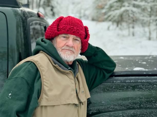 Bearded senior man, resting on his green truck(partial view of truck only),and looking with a serious expression at the camera. Heavily snowed on rural road dirt driveway and snowy woods in the background. The man is wearing a green shirt and tan vest and a homemade red knitted winter hat. Taken in North Wisconsin in Marinette County.