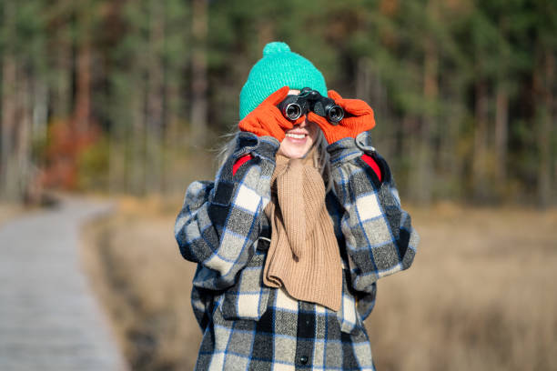 Contented female tourist looking trough binoculars smiling watching wildlife while walking in autumn forest. Joyful woman bird-watcher rest on fresh air, hiking in fall woodland wearing warm clothes.