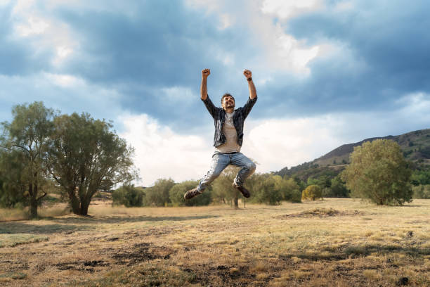Young man leaping with arms raised in victory pose against a backdrop of mountains and cloudy sky. Concept of freedom, success, and overcoming challenges in nature.