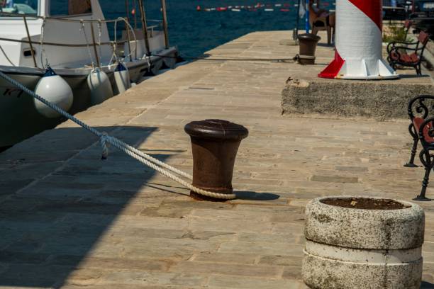 A harbor dock featuring a mooring bollard with a tied rope, benches, and a red-and-white striped lighthouse beside anchored boats.