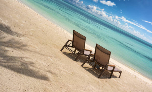 A miniature beach sun lounger with purple and white stripes rests on sunlit sand. A miniature striped beach chair, casting a shadow beside a blurry backdrop. Relaxation and leisure on summer vacation.