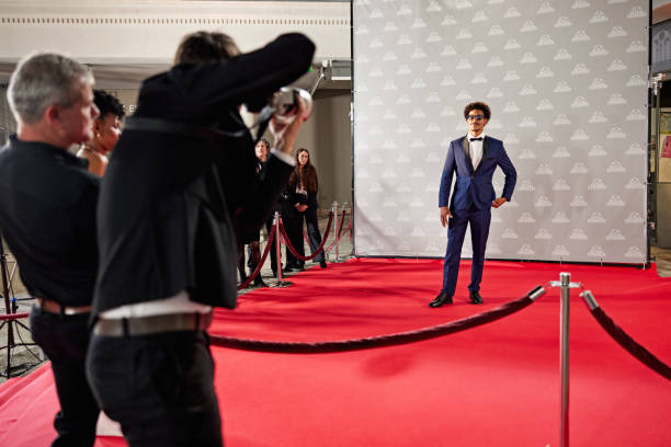 View from behind roped stanchions as professionals document young Black man in blue tuxedo standing against step and repeat wall.