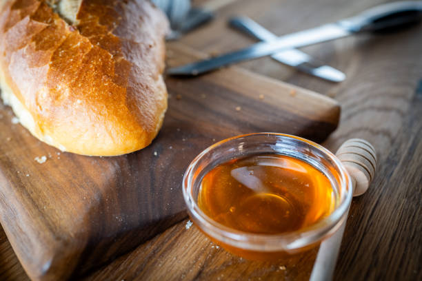 A close-up image of freshly baked bread next to a small bowl of honey and a honey dipper, placed on a wooden cutting board, with utensils slightly blurred in the background.