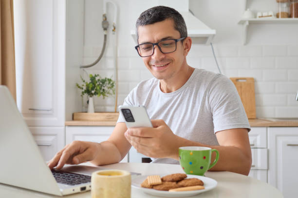 istock Smiling adult businessman using laptop and smartphone for remote work being happy and while browsing social media and managing projects sitting alone in kitchen 2171134945