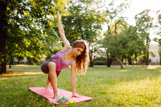 A woman practices yoga in a sunlit park while using a mat and weights on a warm afternoon.