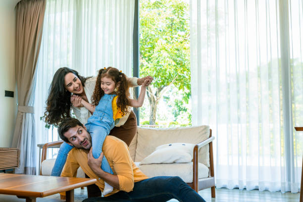 istock The mother sits on the sofa, smiling warmly as she interacts with her daughter, who is perched atop her father’s shoulders. The scene captures a moment of togetherness, contentment, and shared joy in the family’s everyday life. 2171162522
