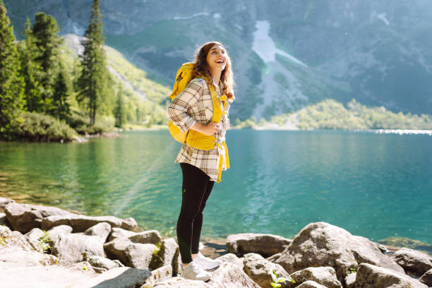istock A young woman with a backpack smiles while posing on rocky terrain beside a clear lake. 2171163921