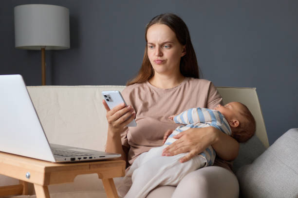 istock Sad disappointed mother using her smartphone to check messages and browse social media while sitting on the couch with her child and working remotely on laptop 2171171206