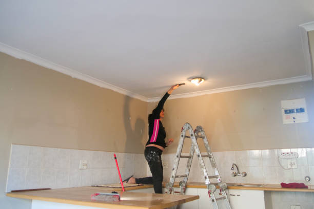 Woman standing on a kitchen countertop and sanding a ceiling while doing home improvements. She is working inside an apartment doing DIY work.