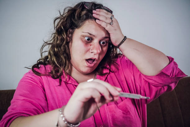 young woman  sitting on the bed and holding her head while holding a thermometer and measuring her temperature