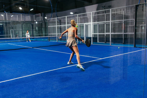 Blonde female padel player mid-motion during a padel match on a brightly lit indoor padel court with blue surface.