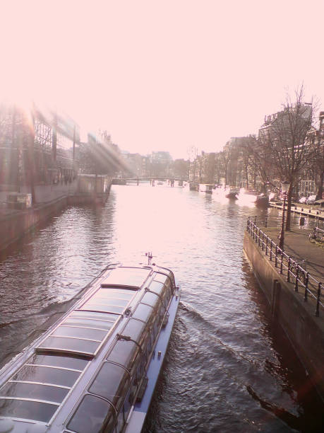 This image showcases a canal scene bathed in sunlight, with a boat, possibly a tourist cruise, prominently in the foreground. The waterway is lined with trees and buildings, and a gentle curve of the canal draws the viewer's eye into the distance where a bridge can be seen. The bright sunlight, likely from a low afternoon sun, creates strong highlights, lens flares, and reflections off the water, adding a sense of warmth and glow to the scene. The sunlight partially obscures the left side of the image, giving the entire composition a dreamy and ethereal quality. The boat, with its glass-covered cabin, seems to be moving along the canal, evoking a sense of peaceful travel through a classic European city. The tranquil atmosphere is enhanced by the reflection of the buildings and light on the water, capturing the essence of a serene canal-side experience.