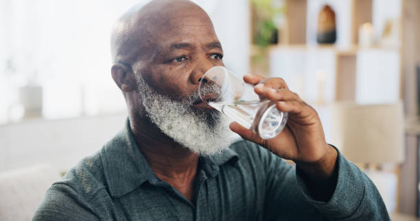 istock Drinking water, mature man and hydration in home, lounge and thinking for wellness, liquid and glass. Living room, healthy and fluid for digestion, thirsty and nutrition in house and black person 2171203314