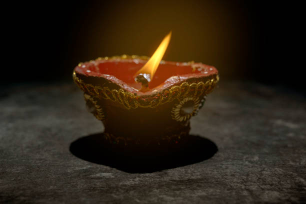 A red and gold decorated pot with a blue and white design sits on a dark surface. A small flame is burning inside the pot. Diwali concept