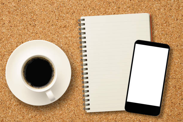 Overhead shot of a cup of coffee on cork table with blank smartphone and blank spiral notebook.