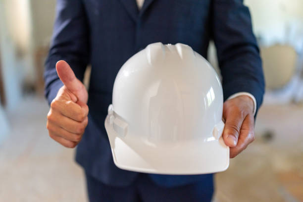 An enthusiastic architect in a smart suit holds a white safety helmet, giving a thumbs up. This image reflects professionalism and commitment to safety in house remodeling and construction