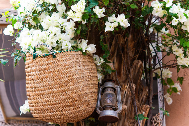 A wicker basket hanging from a palm tree trunk, with a plant with beautiful white flowers. A metal lantern is placed next to it. Horizontal image with an area for copy space