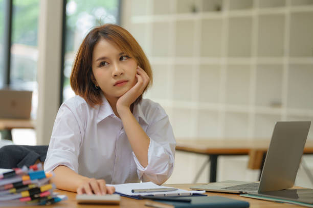 istock Image of a young beautiful brooding Asian woman working with a laptop while sitting at a computer in the office, thinking of professional plans, project management, and considering new business ideas. 2171298837
