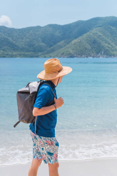istock Man with straw hat and backpack walking along the beach with mountains in the background 2171301356