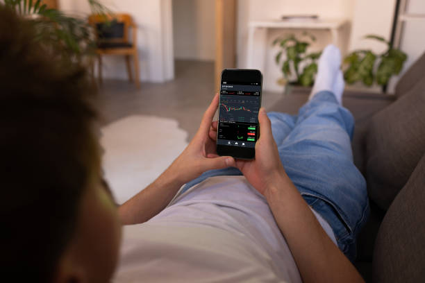 Analysing the Stock Market A high-angle over-the-shoulder view of an unrecognisable young man using a smartphone, he is reading financial stock market analysis while lying down on the sofa in a home in Newcastle, England. man looking at phone over shoulder stock pictures, royalty-free photos & images