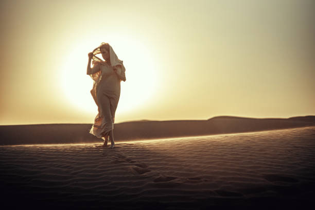 Woman in flowing dress silhouetted against radiant sunset in desert. Ethereal atmosphere as she lifts fabric. Embodies freedom, spirituality, and connection with nature. Ideal for lifestyle and travel themes. mujer con vestido fluido recortada contra la radiante puesta de sol en el desierto. ambiente etéreo mientras levanta la tela. encarna la libertad, la espiritualidad y la conexión con la naturaleza. ideal para temas de estilo de vida y viajes.