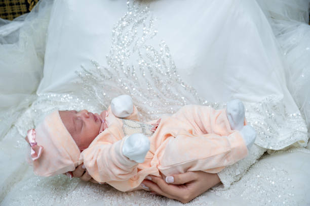 A heartwarming top view of a newborn baby girl swaddled in a soft peach blanket, resting on the intricate lace details of a white wedding dress. This image beautifully captures the concept of new beginnings and family traditions, ideal for use in family oriented content, celebrations, and lifestyle articles