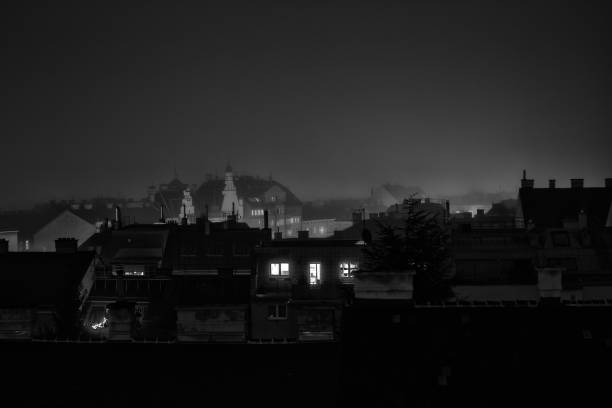 A moody black and white photograph of a city skyline at night, with illuminated windows and silhouetted buildings against a foggy background.
