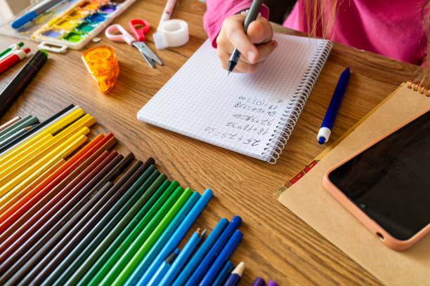 istock Child doing math test in notebook using smartphone with stationery on wooden table. Girl hand writing mathematical exercises as school homework. Learning, education using digital tools concept 2171352061