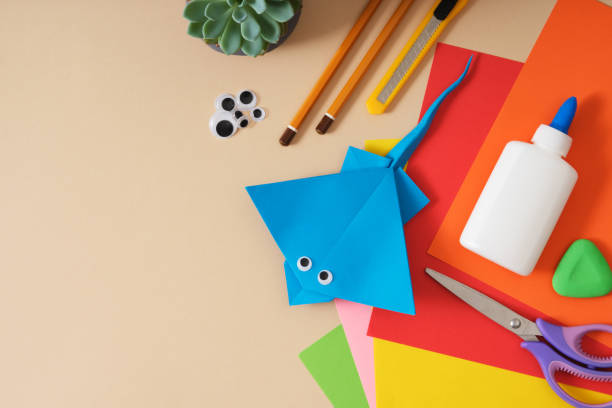 istock Work table for paper crafts colored sheets, glue, scissors, pencils, stationery knife. Finished origami toy stingray. Beige background, space for your text. 2171353778