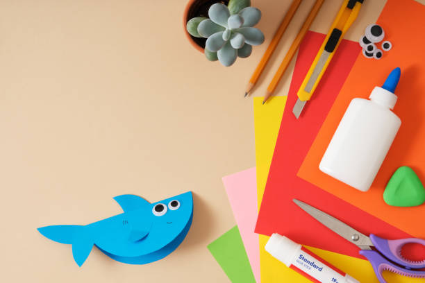 istock Finished paper toy shark. Beige background, space for text. Work table for paper crafts colored sheets, glue, scissors, pencils, stationery knife. 2171353854