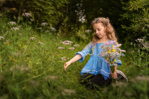 A young girl with curly blonde hair kneels in a meadow, picking wildflowers. She wears a blue blouse and holds a bouquet of colorful flowers.