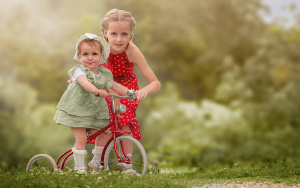 Two young girls outdoors, one older and one younger, sharing a playful moment with a red tricycle. The older girl in a red polka dot dress is helping her younger sibling, dressed in a light green outfit and bonnet, to ride the tricycle. The setting is a lush, green garden with soft, natural light, creating a nostalgic and carefree atmosphere.