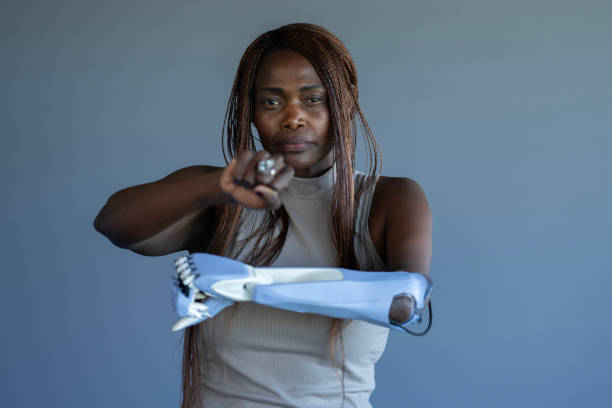 A female arm amputee of African decent poses in-studio for a portrait.  She is dressed casually and smiling gently as she expresses her resilience and strength while wearing her prothesis.