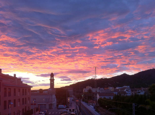 The image captures a beautiful sunset (or sunrise) over a small town or village. The sky is filled with vibrant shades of pink, orange, and purple, creating a stunning contrast with the darker silhouettes of the town's buildings and the surrounding hills. The clouds in the sky add texture and depth, reflecting the warm colors of the sun.\n\nIn the foreground, there are buildings and streets with a few cars and streetlights, while in the background, a prominent church tower rises against the colorful sky. The hills or mountains behind the town provide a picturesque backdrop, blending natural beauty with the charm of a peaceful town.