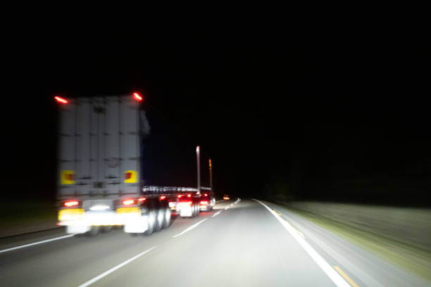 The view from the front windshield of a car traveling along a dark highway at night, overtaking large trucks as the road stretches ahead. The headlights illuminate the asphalt, casting long shadows and reflecting off the surrounding trucks as the car smoothly passes them.