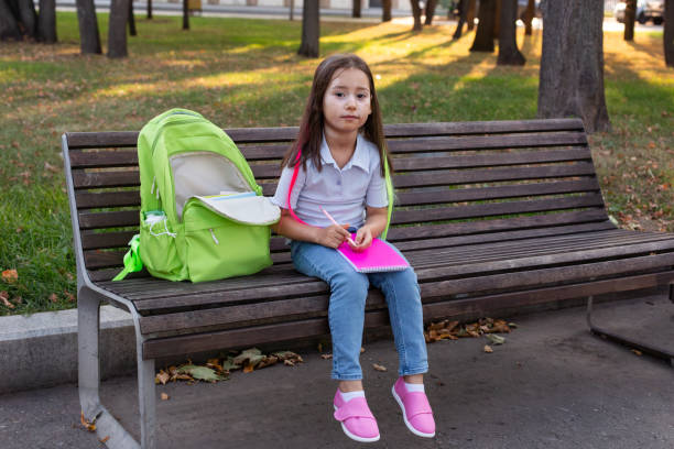 istock Young girl with backpack sitting on bench writing in notebook outdoors 2171444254