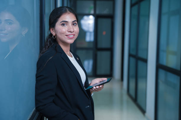 istock Young indian female corporate employee standing in office hallway looking at camera while holding mobile in hand. workplace and corporate culture concept. 2171459510