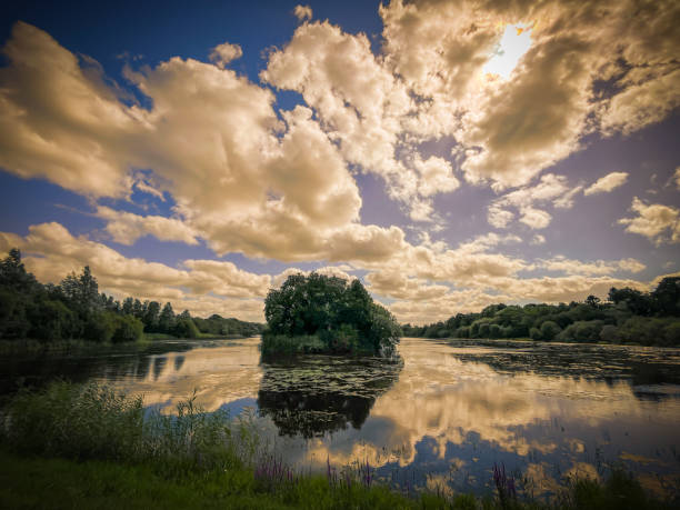 A serene view of Rinn Lough in County Leitrim, where trees and clouds are reflected on the calm waters. The landscape is framed by lush greenery and a sky filled with soft, billowing clouds.