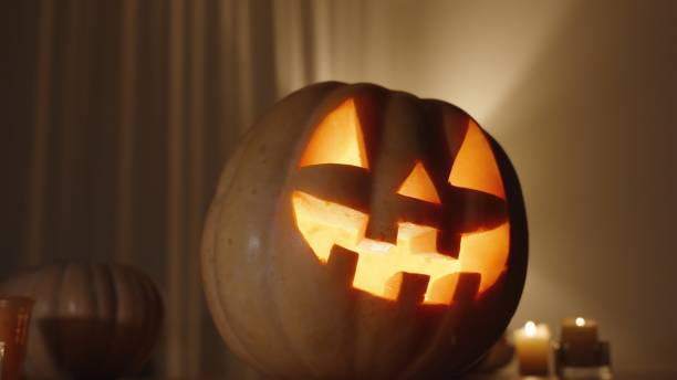 istock Pumpkin with a Carved Halloween Jack-o'-Lantern Symbol, Fire Burning Inside It, in a Dark Room on the Table with Pumpkins and Candles. Warm Tones of Home Interior. 2171494212