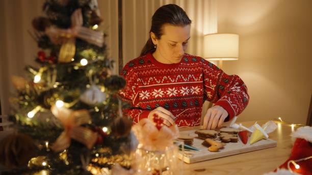 Focused woman in a red festive sweater decorates gingerbread cookies by a lit Christmas tree, embodying the holiday spirit.